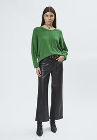 Grüner gestrickter Pullover mit rundem Ausschnitt und strukturiertem Vorderteil, kombiniert mit schwarzen, weiten, aus Kunstleder gefertigten Hosen und Absatzstiefeletten.