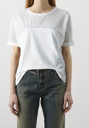 Kobieta w białej koszulce z krótkim rękawem z przezroczystym górnym panelem i ciemnych, wysokich dżinsach stojąca na jednolitym tle.