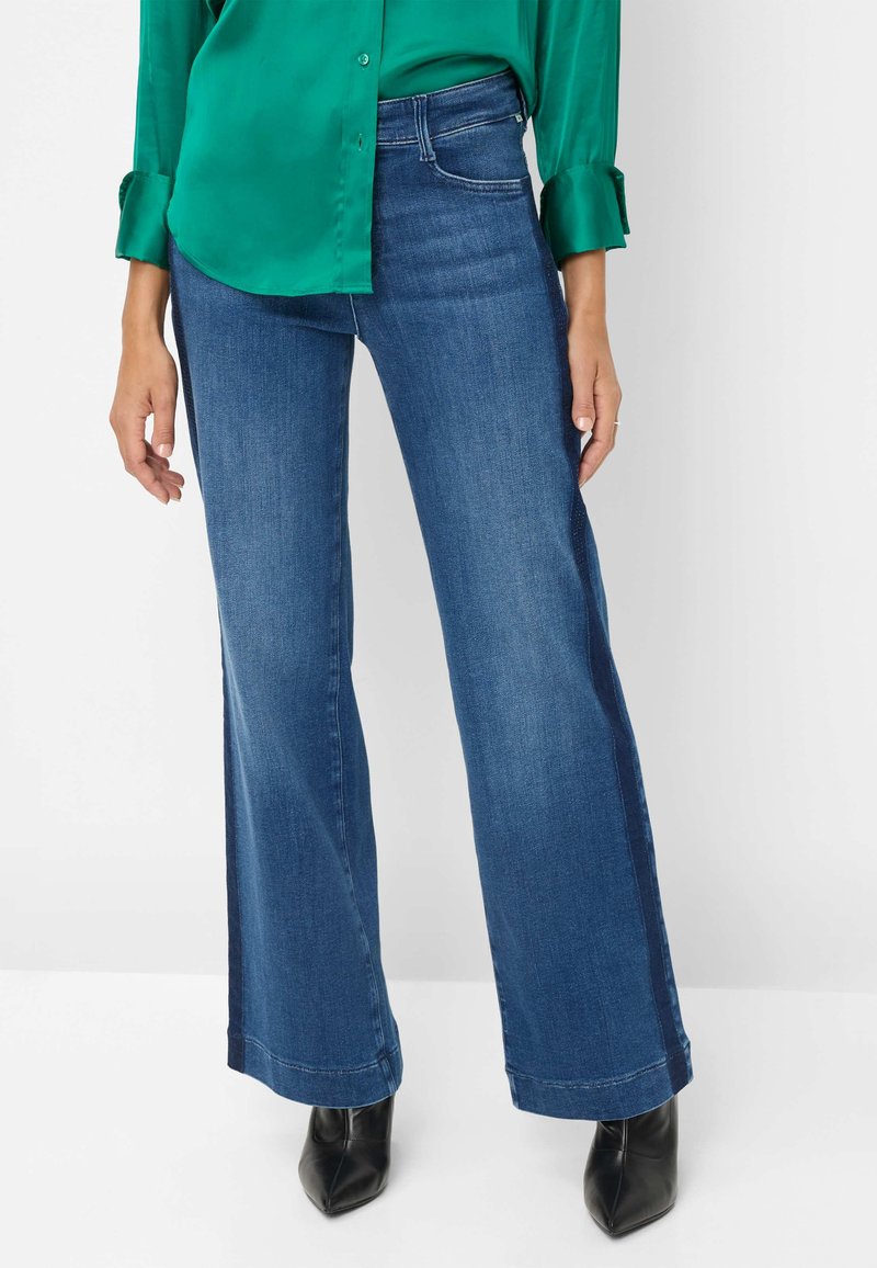 Wijdvallende jeans van donkerblauwe denim. Heeft een middelhoge taille, een klassieke vijf-pocket stijl en een gestikte zoom. Gecombineerd met een groene satijnen blouse.