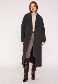 Cappotto lungo doppiopetto in lana grigia scura con ampi rever e bottoni dorati; abbinato a un maglione leggero in maglia e pantaloni di velluto a coste marroni.