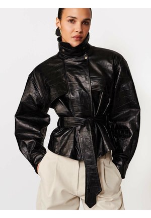 Naine kannab läikivat musta krokodillinahast jope, millel on kõrge krae, vööga talje ja kreamvalged püksid.