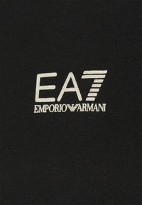 La tela negra presenta un destacado logo plateado "EA7 EMPORIO ARMANI" con elementos estilizados, exhibiendo una textura suave y un diseño limpio.