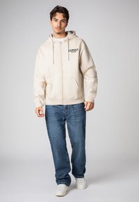 Beige Kapuzenpullover mit Reißverschluss, einer Fronttasche und dem schwarzen Schriftzug "EXPECT NOTHING". Kombiniert mit blauen Jeans und weißen Turnschuhen. Glatte Textur.
