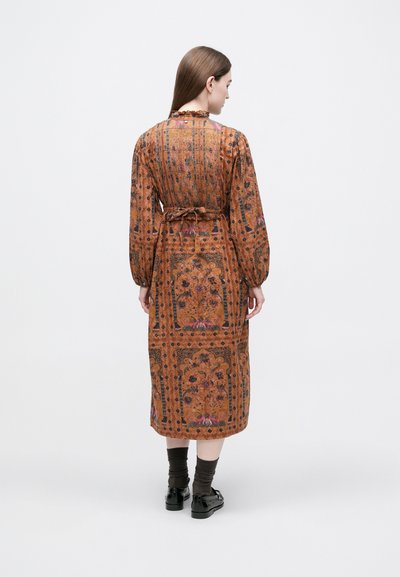 Moteris su ilgais ruda plaukais, dėvinti rudos spalvos midi suknelę su raštais, ilgomis rankovėmis, susirištu diržu, tamsiomis kojinėmis ir juodais batais, stovinti nugara.