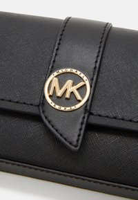 Cartera de cuero texturizado negro que presenta un logo circular dorado con "MK" y "MICHAEL KORS" grabados, asegurada con un cierre a presión.