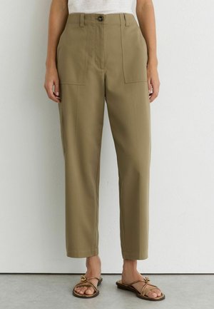 Femme portant un pantalon kaki taille haute, jambes droites, avec des poches plaquées à l'avant et des sandales plates marron à lanières, debout contre un mur blanc.