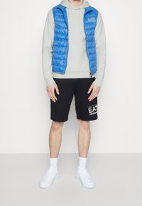 Uomo che indossa un hoodie grigio chiaro, un gilet trapuntato blu, pantaloni corti neri con logo bianco, calze bianche e sneaker bianche, in piedi contro uno sfondo semplice.