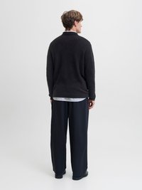 Uomo con maglione nero, camicia a righe e pantaloni scuri larghi, in piedi di fronte a uno sfondo bianco semplice, visto da dietro.