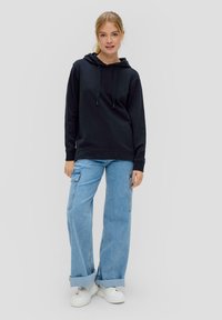 Zwarte hoodie met trekkoorden, gecombineerd met lichtblauwe wijde jeans en witte sneakers. Glad materiaal en casual ontwerp.