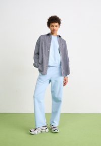 Sweatshirt bleu à col rond, chemise à rayures sous un pull, et jogging bleu clair associés à des baskets à motifs sur un sol vert.