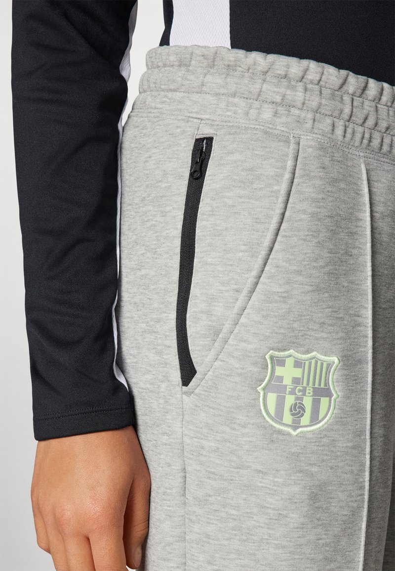 Primo piano di pantaloni della tuta grigi con tasca con zip nera ed emblema del FC Barcelona, indossati con un top a maniche lunghe nero.