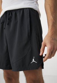 Homme portant un short de sport noir avec un logo Jordan blanc près de l'ourlet et un débardeur à côtes blanches, main visible avec un tatouage au poignet.