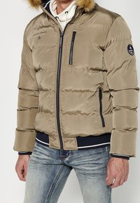 Homme portant une doudoune beige avec capuche bordée de fourrure, un sweat à capuche blanc en dessous et un jean bleu, debout avec les mains détendues le long du corps.