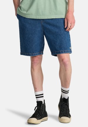 Persona che indossa shorts di denim blu, calzini bianchi con strisce nere, sneakers nere alte e una maglietta verde con le mani nelle tasche.
