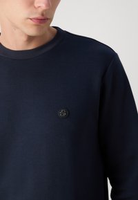 Mörkblå sweatshirt med rund halsringning och subtil textur. Har en svart cirkulär logotyp på bröstet, tillverkad av mjukt tyg.