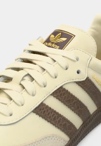 Buty sportowe Adidas Samba w kremowej skórze z brązowymi paskami zamszowymi. Posiadają okrągłe sznurowadła, złote logo oraz fakturowane detale na paskach.