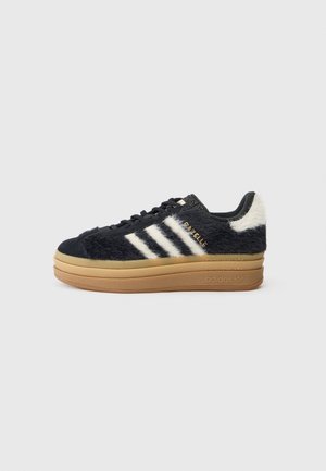 Baskets Adidas noir et blanc avec texture duveteuse, trois bandes blanches sur le côté, semelle épaisse couleur marron clair et lacets noirs sur fond uni.