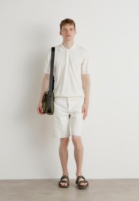 Polo shirt blanc, shorts blancs et sandales marron. Portant un sac bandoulière vert. Design simple et couleurs claires, look minimaliste.