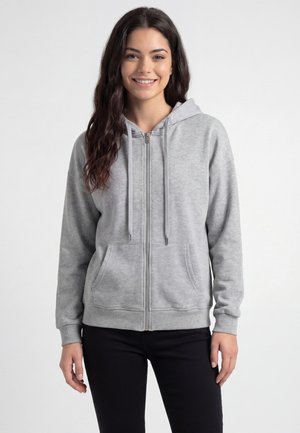 Young woman with long dark hair wearing a light gray zip-up hoodie and black pants, smiling against a plain white background.