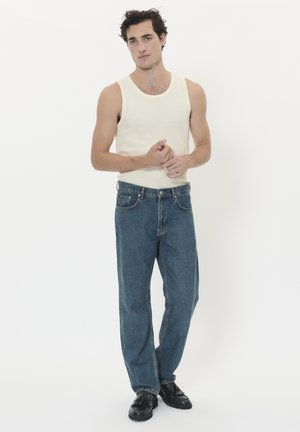 Camiseta sin mangas beige claro y vaqueros azules de corte holgado, con bolsillos estándar y zapatos negros, sobre un fondo blanco liso.