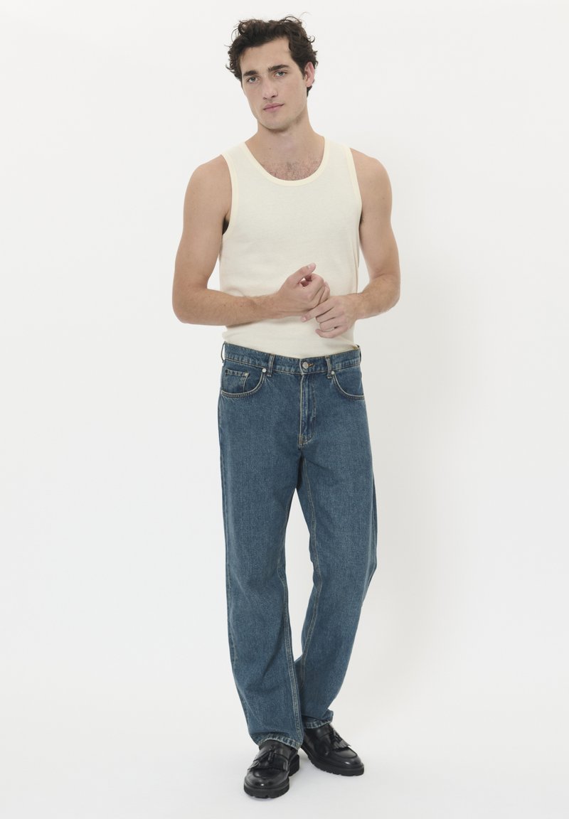 Šviesiai smėlio spalvos rankovių marškinėliai ir laisvai prigludusios mėlynos džinsinės kelnės, baigtos standartinėmis kišenėmis ir juodais batais, fone paprasta balta spalva.