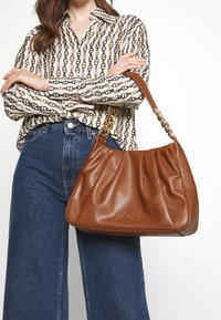 Borsa a spalla in pelle marrone con una design arricciato e tracolla a catena. Esibita su una camicia stampata e jeans a vita alta.