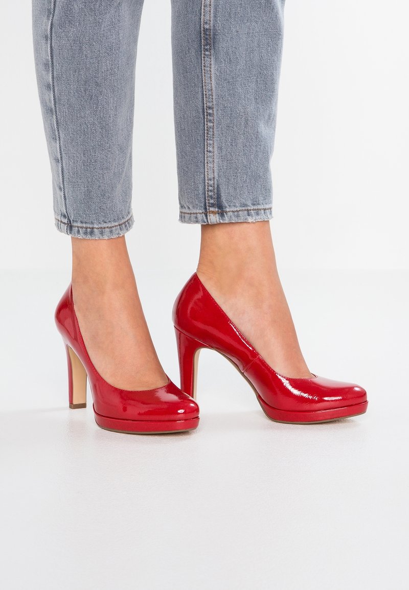 Femme portant un jean bleu clair et des escarpins rouges brillants, debout sur un sol blanc.
