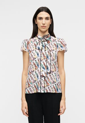 Γυναίκα με μακριά σκούρα μαλλιά, που φοράει μπλούζα με κοντά μανίκια και πολύχρωμες αφηρημένες φιγούρες, με δεμένο φιόγκο στο γιακά, συνδυασμένη με μαύρο παντελόνι.