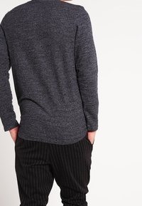 Suéter de punto gris oscuro con mangas largas y un dobladillo curvado, combinado con pantalones rayados negros. Presenta un material texturizado.