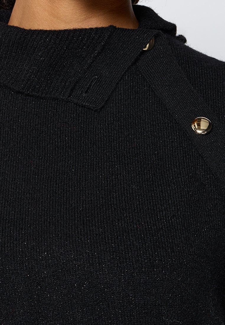 Dettaglio ravvicinato di un maglione nero a maglia con un colletto alto e due bottoni a pressione dorati sulla spalla.