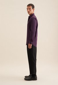 Camicia lunga viola con colletto button-down, abbinata a pantaloni e scarpe neri. La camicia presenta un tessuto morbido e una vestibilità su misura.