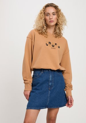 Jasnobrązowy sweatshirt z nadrukiem tekstu "CROSS JEANS" w kolorze czarnym i złotym, zestawiony z ciemnoniebieską spódnicą dżinsową do kolan.