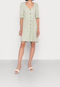 Kobieta w jasnozielonej, fakturowanej sukience na guziki z bufkowymi rękawami i białych, plecionych sandałach na obcasie, stojąca na gładkiej podłodze.
