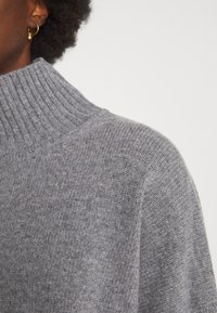 Grijze gebreide trui met een hoge ribbels halslijn, zachte textuur en een ontspannen pasvorm. De stof heeft een lichte gemêleerde patroond.