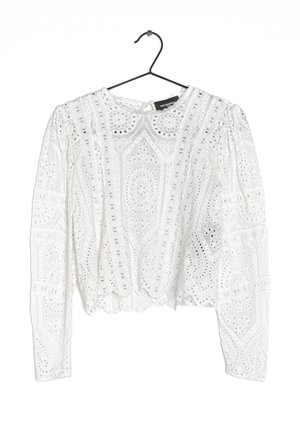 Blouse blanche à manches longues en dentelle avec des motifs de broderie floraux et géométriques, suspendue à un cintre noir sur un fond blanc.