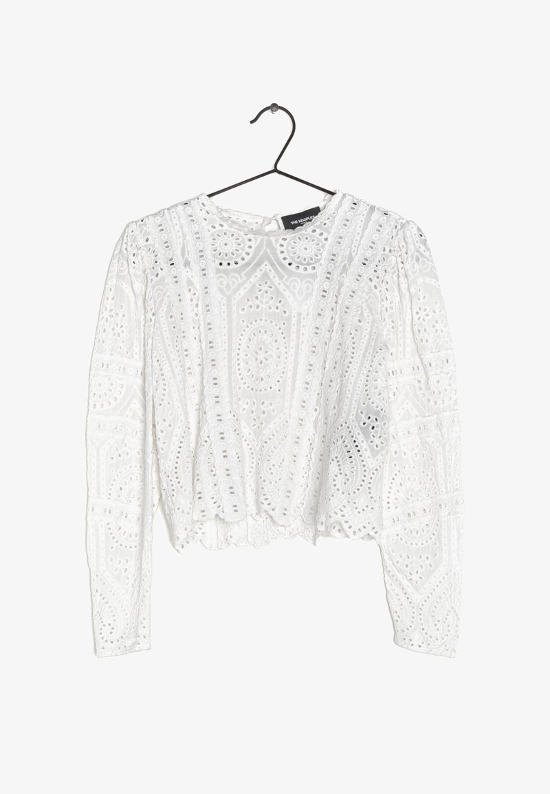 Blouse blanche à manches longues en dentelle avec des motifs de broderie floraux et géométriques, suspendue à un cintre noir sur un fond blanc.