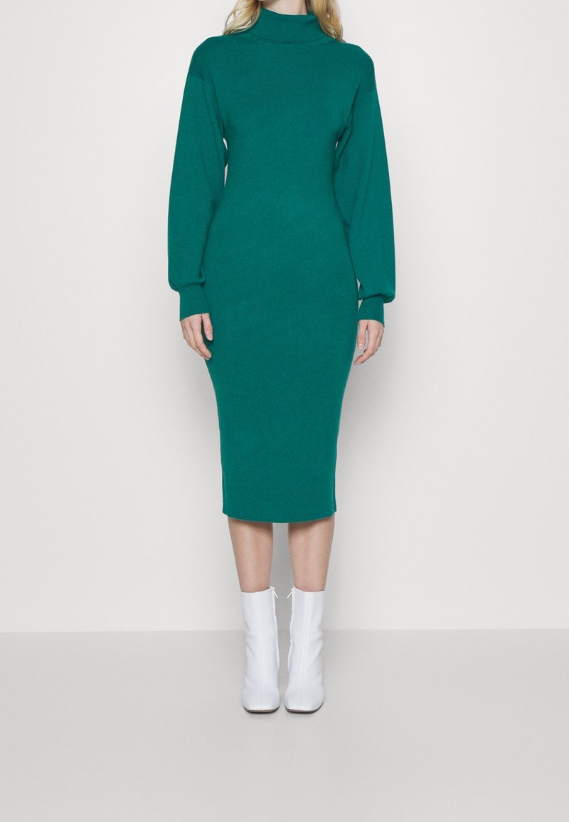Femme portant une robe midi ajustée à manches longues de couleur bleu-vert avec un col montant et des bottes blanches à la cheville, se tenant devant un fond uni.