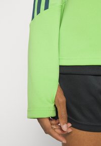 Zielona sportowa kurtka z długimi rękawami, z granatowymi paskami na ramieniu i dopasowanym krojem. U dołu widoczne czarne spodenki.