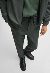 Gray patterned trousers made of textured fabric paired with a green ribbed sweater and black slip-on shoes, featuring a drawstring waist.