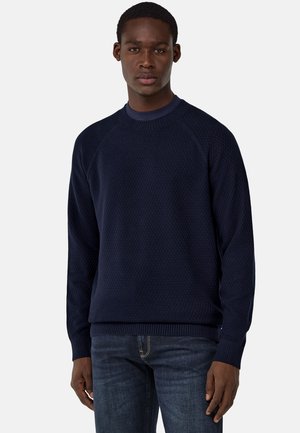 Dunkelblauer Strickpullover mit strukturiertem Muster, Rundhalsausschnitt und gerippten Bündchen. Kombiniert mit dunklen Jeans, die eine lockere Passform zeigen.