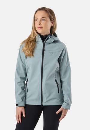 Woman with long blonde hair wearing a light blue zip-up hooded jacket and black pants, looking slightly to the left.