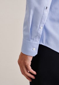 Camicia a scacchi blu chiaro con polsini abbottonati; presenta bottoni bianchi e un dettaglio ricamato di rosa nera sul polsino. Tessuto morbido e testurizzato.