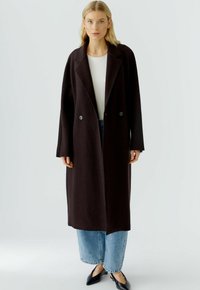 Lang, donkerbruin wollen coat met een losse pasvorm, brede revers en dubbele knopen. Gecombineerd met lichte blauwe wijd uitlopende jeans en zwarte spitse schoenen.
