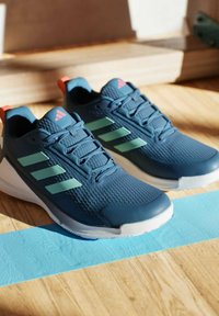 Un paio di scarpe da corsa Adidas blu navy con strisce verde chiaro e suole bianche posate su un pavimento di legno con una linea di nastro blu.