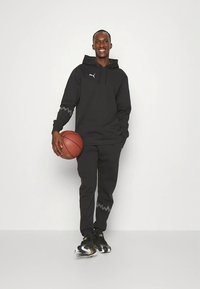 Sweat à capuche athlétique noir avec une poche avant et des accents en zigzag gris ; pantalon noir assorti ; baskets noires et blanches ; tenant un ballon de basket.