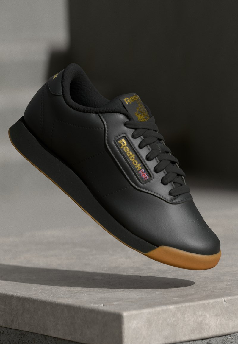 Sapatilha de couro preto com detalhes amarelos e logótipo, apresentando uma superfície texturizada, cordões e uma sola de borracha. Leve, com um design aerodinâmico.