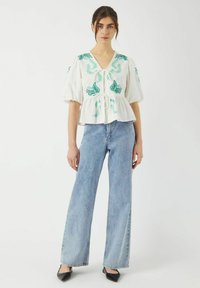 Hvid bluse med grøn broderi, korte puffærmer, V-hals og snor i taljen, parret med lyseblå brede jeans og sorte sko.