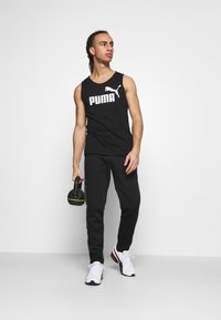 Camisola de alças preta sem mangas com o logo branco "PUMA", calças de treino pretas e sapatilhas brancas com detalhes em vermelho e preto, segurando um kettlebell.