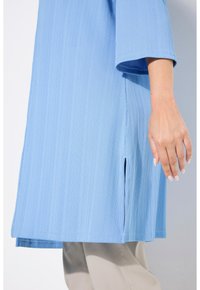 Himmelblaue Tunika mit gerippter Textur und kurzen Ärmeln. Verfügt über seitliche Schlitze am Saum für zusätzliche Bewegungsfreiheit. Wird mit beigen Hosen kombiniert.