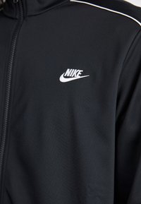 Gros plan sur une veste noire Nike à fermeture éclair avec un logo Nike blanc sur la poitrine et des coutures blanches contrastantes le long des épaules.